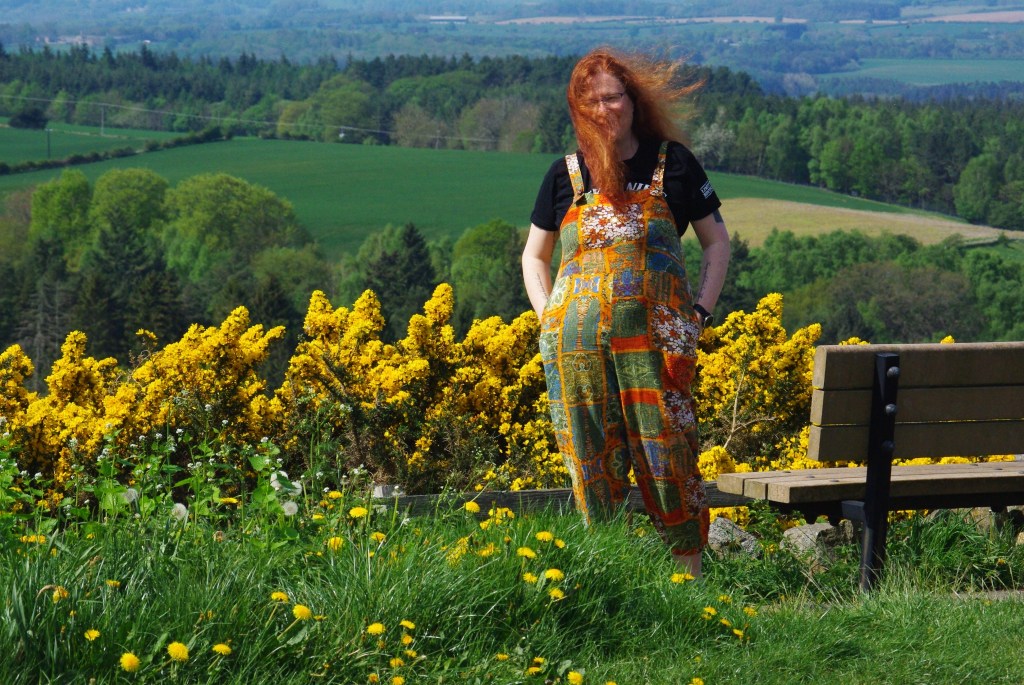 Image shows me, a white person with long red hair, in colourful dungarees on a hillside, next to a bench. Behind me yellow gorse glows.
